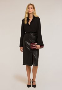 Camicia nera con bottoni e maniche a campana, abbinata a una gonna midi in pelle nera con uno spacco frontale. Tiene una pochette bordeaux.