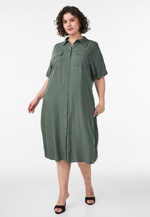 Lühikeste lokkis juustega naine, kes kannab lahtist, nööpidega põlvpikkust oliivrohelist kleiti ja musti avatud varbaga kontsakingi, seisab valge tausta ees.