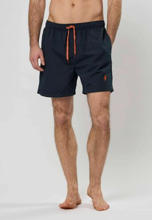 Man met marineblauwe zwemshorts met oranje trekkoord en logo, die op blote voeten staat tegen een neutrale achtergrond.