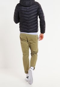 Svart pufferjacka med chevron-sömmar, grå hoodie underneath, olivgröna byxor och vita sneakers med svarta accenter.