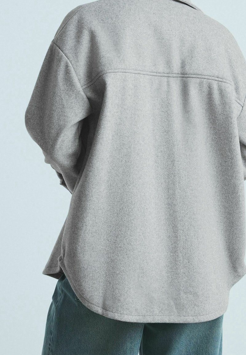 Camicia grigio chiaro, tessuto testurizzato, con orlo curvo e dettagli sulla cucitura posteriore, abbinata a jeans verde scuro. Vestibilità morbida e tessuto morbido.