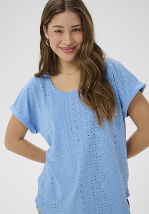 Mladá žena s dlhými hnedými vlasmi, oblečená v modrom krátkym rukávom topu so vzorom, usmievajúca sa s rukami v bok proti jednoduchému svetlému pozadiu.