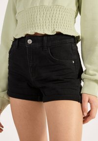 Černé džínové šortky s upraveným střihem, s ohrnutými lemy, předními kapsami a stříbrným knoflíkem. V kombinaci se světle zeleným topem.