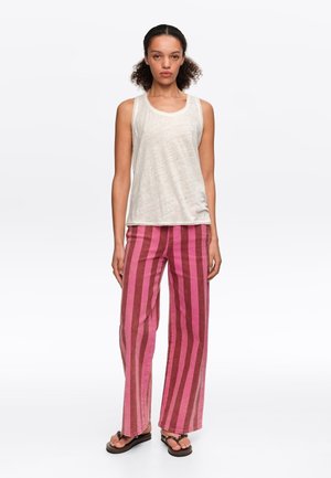 Mujer con top blanco sin mangas, pantalones anchos de rayas verticales rosas y rojas, y sandalias negras, de pie sobre fondo blanco.