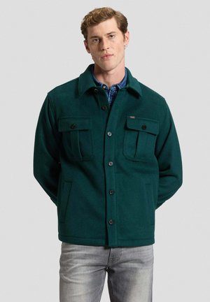 Junger Mann mit kurzen Haaren, der eine dunkelgrüne, geknöpfte Jacke über einem blauen Hemd und hellgrauen Jeans vor einem einfarbigen Hintergrund trägt.