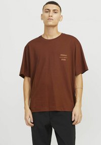 Camiseta de algodón marrón con un ajuste relajado, que presenta un cuello redondo y mangas cortas. Texto impreso pequeño en la parte superior izquierda del pecho.
