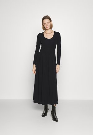 Robe noire à manches longues avec un décolleté rond, corsage ajusté et jupe fluide. Le mannequin porte des bottines noires et un collier en chaîne dorée.