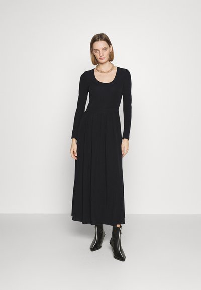 Zwarte jurk met lange mouwen, een ronde halslijn, een aansluitend lijfje en een vloeiende rok. Het model draagt zwarte enkellaarsjes en een gouden ketting.