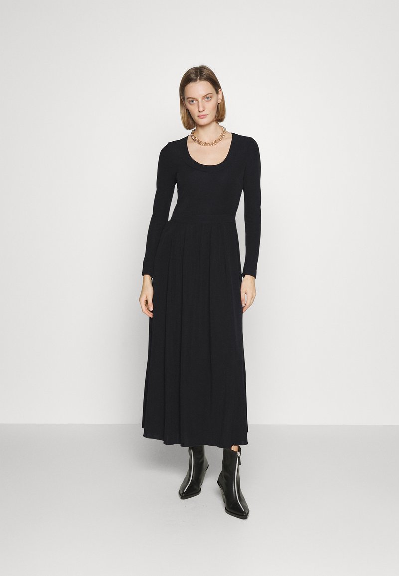 Robe noire à manches longues avec un décolleté rond, corsage ajusté et jupe fluide. Le mannequin porte des bottines noires et un collier en chaîne dorée.