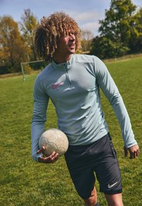 Lyseblå langærmet athletic top med halv-lynlås og pink Nike-logo, parret med mørke shorts, holder en slidtagen fodbold på græs.