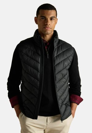 Schwarze gepolsterte Weste mit einem Chevron-gesteppten Design, Reißverschluss vorne und seitlichen Taschen, getragen über einem schwarzen Pullover mit einem bordeauxfarbenen Kragenhemd.