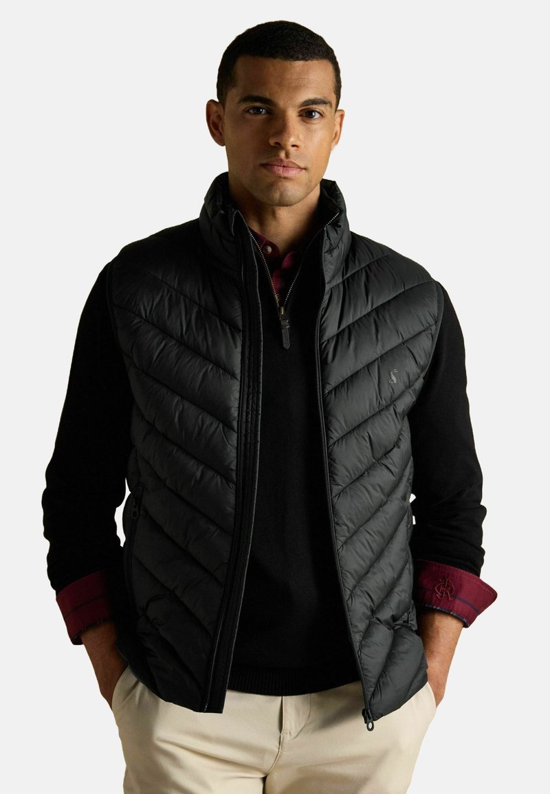 Čierna prešívaná vesta s diamantovým vzorom, so zipsom v prednej časti a bočnými vreckami, nosená na čiernom svetri s vínovou košou.