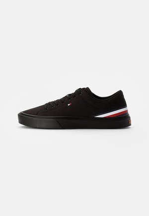 Scarpa da ginnastica nera bassa con suola testurizzata, lacci neri e strisce rosse, bianche e blu sul tallone e sul lato esterno.
