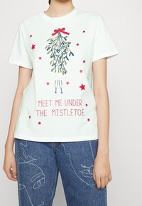 Vit bomullströja med en färgglad mistelgrafik, röda stjärnor och texten "MÖT MIG UNDER MISTELN" i rött.