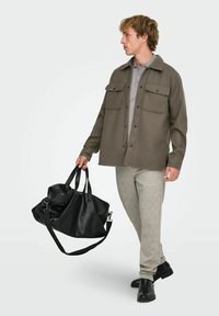 Chemise-veston boutonnée vert olive avec deux poches poitrine, polo gris, pantalon gris clair, sac de voyage en cuir noir et chaussures noires.