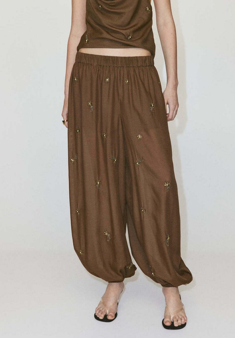 Pantalon ample marron avec petite broderie florale, taille élastique et poignets froncés, porté avec des sandales à brides transparentes sur un mannequin debout.