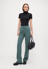 Musthave mustriba lühikeste varrukatega kõrge kaelusega särk koos teal värvi elastse vöökohtadega laia jalaga pükstega. Mudel hoiab käes väikest musta käekotti. Mustad jalanõud.