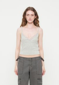 Jeune femme aux longs cheveux ondulés portant un caraco gris clair orné de dentelle et un pantalon cargo gris foncé sur fond blanc.