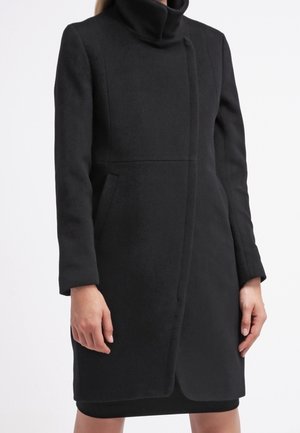 Femme portant un manteau noir longueur genou avec un col montant et des manches longues, debout les bras détendus le long du corps.