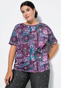 Tričko s krátkým rukávem s živým patchworkovým designem v purpurové, modro-zelené a černé barvě, zahrnujícím květinové a geometrické vzory; nošeno s černými flitrovými kalhotami.