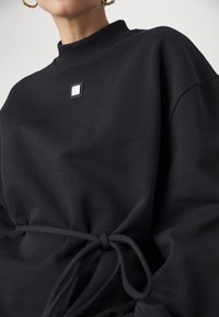 Haut noir à manches longues avec col haut côtelé et lien à la taille, porté par une personne portant des boucles d’oreilles créoles dorées.