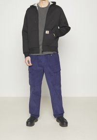 Sudadera negra con cremallera y forro gris, dos bolsillos frontales; combinada con pantalones cargo morados con bolsillos laterales y zapatillas negras.