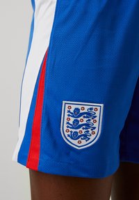 Pantalones cortos deportivos azules con acentos laterales blancos y rojos, que cuentan con una tela texturizada y un escudo con tres leones y detalles florales.