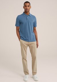 Modra polo majica iz mehke bombažne tkanine, kratki rokavi, klasičen ovratnik, v kombinaciji s svetlim bež chinos hlačami in belimi supergami, stoji na nevtralni podlagi.
