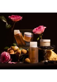 Drie huidverzorgingsproducten van OUAI: een doorzichtige fles met het label "Detox Shampoo", een wit potje en een doos met gouden accenten, omringd door roze rozen, fruit en een zwarte achtergrond.