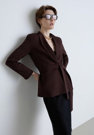 Personne portant un blazer marron foncé ceinturé et une jupe noire, accessoirisée de grandes lunettes de soleil et d'un collier chaîne en or, appuyée contre un mur blanc.