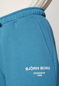 Tyrkysové jogger nohavice vyrobené z mäkkej tkaniny, s elastickým pásom a bočnou kapsou. Biele logo s nápisom "BJÖRN BORG STOCKHOLM 1956."