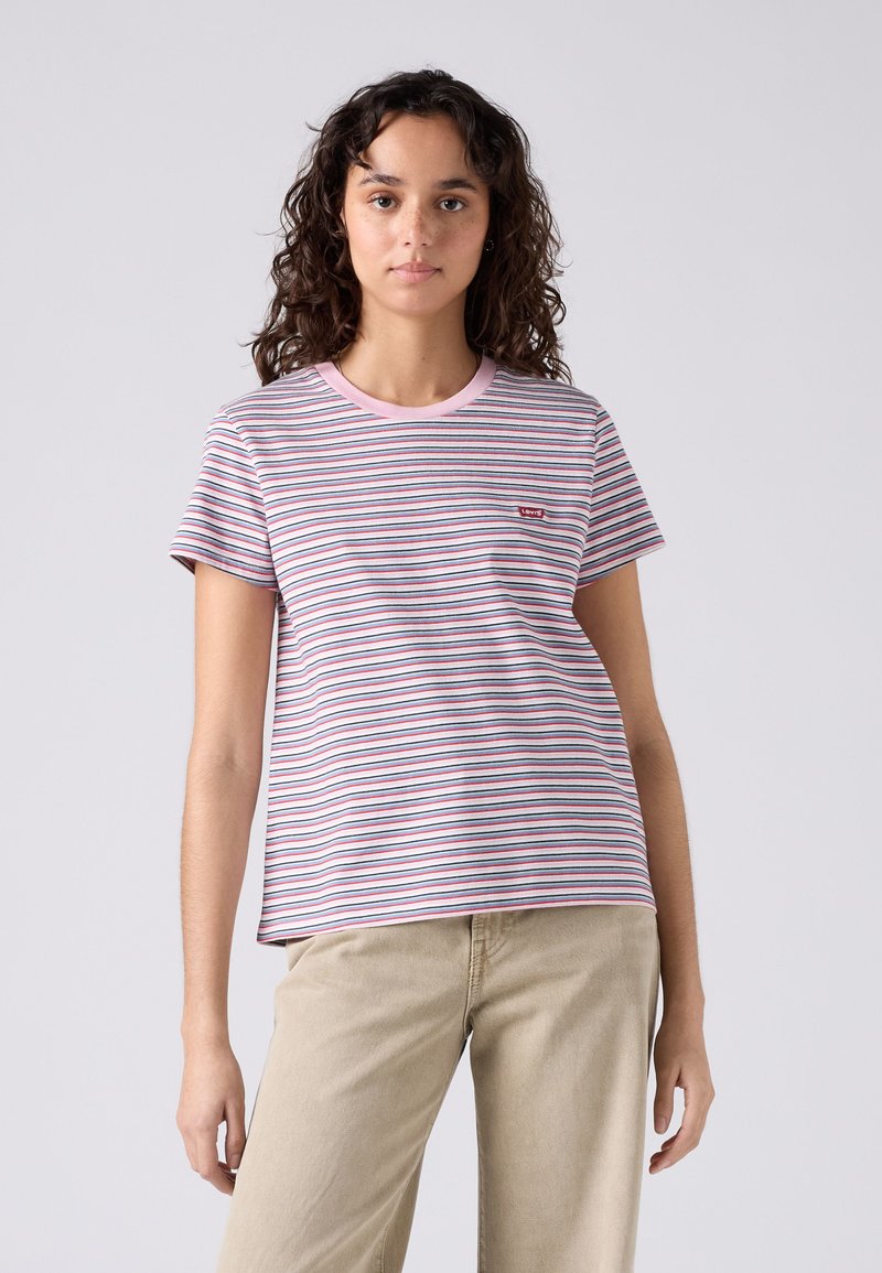 Mujer joven con cabello rizado que lleva una camiseta de rayas rosas y blancas y pantalones beige, de pie frente a un fondo claro y sencillo.