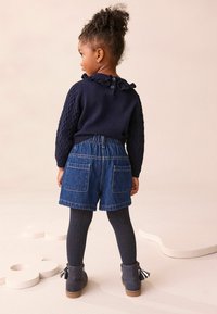 Marineblå strikket sweater med flæsekrave, denimshorts og marineblå ribbede strømpebukser. Brune støvler med frynse-detalje. Bagview.