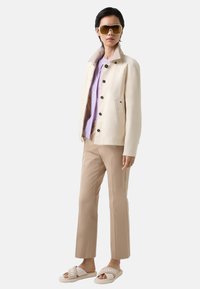 Beige Jacke mit strukturiertem Kragen, lavendelfarbenes Hemd, beige Hose und weiße Slides mit Logo-Text. Model posiert vor einem einfarbigen Hintergrund.