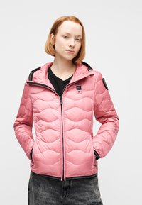 Νέα γυναίκα με κοντά κόκκινα μαλλιά φοράει ροζ κοντή μπουφάν με ραφές πάνω από μαύρο πουκάμισο και σκούρα τζιν, με τα χέρια στις τσέπες του μπουφάν.