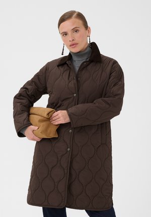 Donna con un lungo cappotto trapuntato marrone e dolcevita grigio, che tiene in mano una pochette piegata in camoscio color cognac su sfondo neutro.