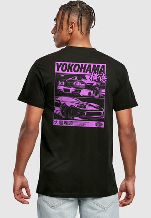 Jeune homme aux cheveux tressés portant un T-shirt noir avec un graphisme violet de deux voitures de sport et le texte "YOKOHAMA" dans le dos.