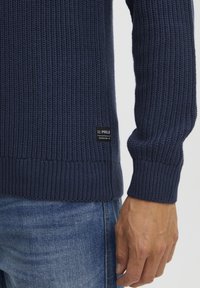 Marineblauer Strickpullover mit gerippter Textur, ausgestattet mit einem Logo-Patch an der unteren Seite und enganliegenden Bündchen. Kombiniert mit Jeans.