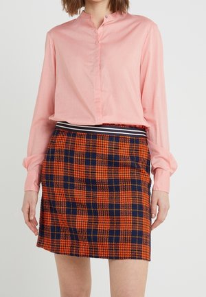 Mujer con blusa de manga larga rosa claro y falda mini de cuadros naranja y azul marino con cinturilla de rayas, de pie frente a un fondo liso.