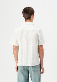Camisa blanca de manga corta con un patrón texturizado que presenta cortes verticales. Diseño con cuello, dobladillo recto y aberturas laterales.