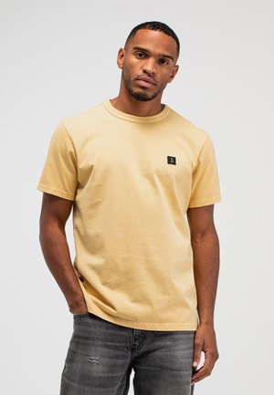 Man draagt lichtgeel kortemouw T-shirt met zwarte patch op de borst en zwarte versleten spijkerbroek, poserend met één hand in de zak.