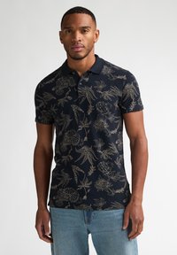 Mørkeblå polo med korte ærmer, der har et gyldent blomsterprint og en krave. Bæres med lyseblå denimjeans.