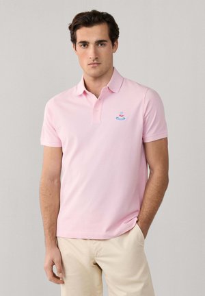 Hombre joven con una camisa polo de manga corta color rosa claro y pantalones beige, de pie con una mano en el bolsillo contra un fondo liso.