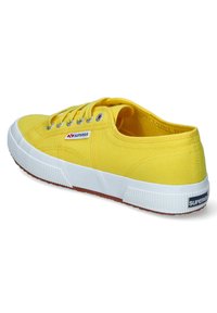 Sneaker in tela gialla con suola in gomma, dettagli bianchi, occhielli argentati e un piccolo logo rosso e blu sul lato. Parte superiore testurizzata.