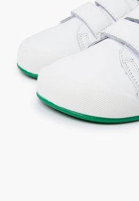 Zapatos blancos con una superficie texturizada, correas de velcro y suelas de goma verdes contrastantes. Diseño de punta redondeada y líneas simples y limpias.