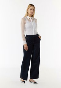 Weißes durchscheinendes Hemd mit Knöpfen und zwei Brusttaschen, kombiniert mit hoch taillierten, dunkelblauen, weiten Jeanshosen mit goldenen Akzenten.