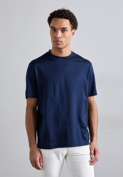 T-shirt bleu marine à manches courtes avec un motif texturé subtil, col rond et coupe décontractée, associé à un pantalon blanc.
