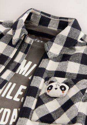 Fleecejacke mit schwarz-weißem Karomuster, Reißverschluss und Panda-förmiger Verzierung auf der Tasche. Graues T-Shirt mit sichtbarem Aufdruck darunter.