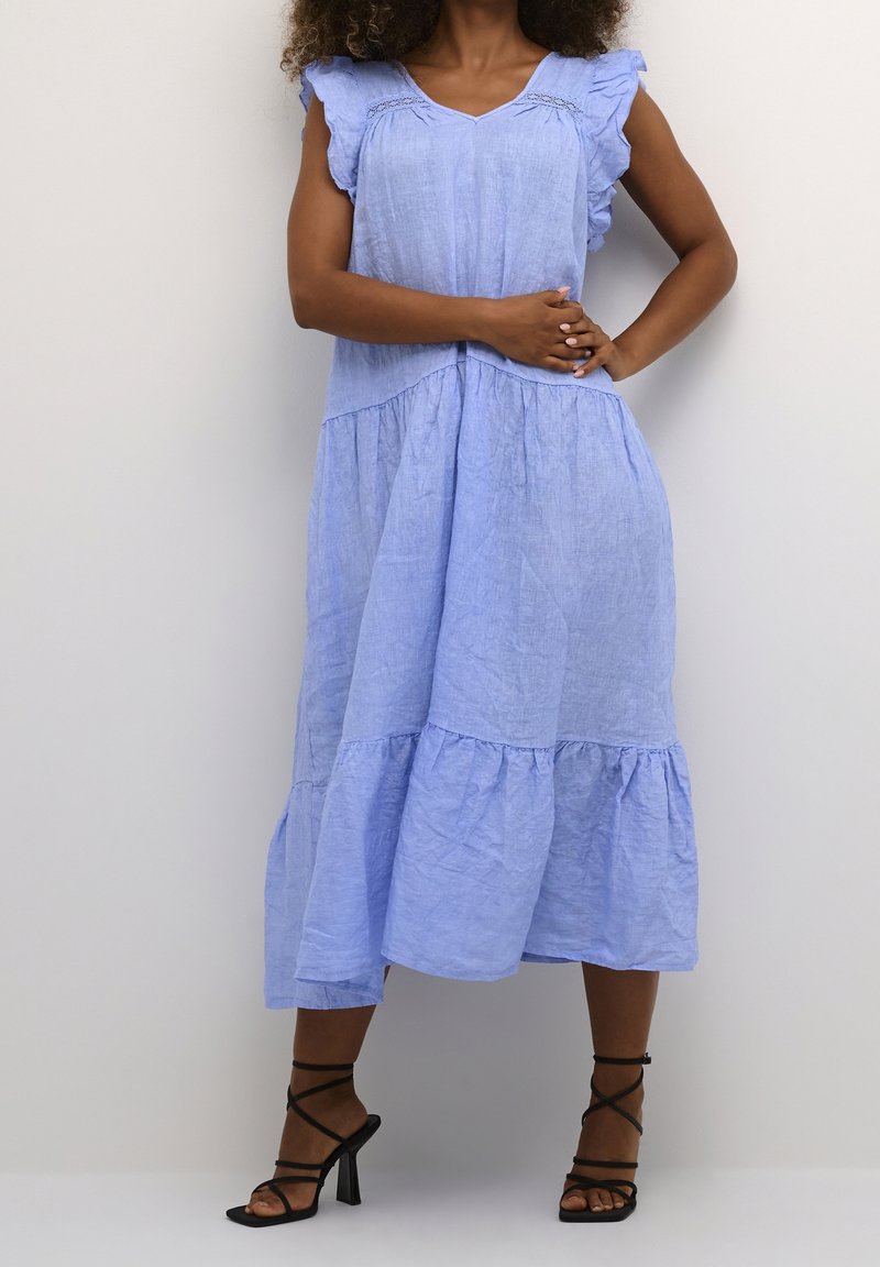 Femme portant une robe midi bleu clair sans manches avec des détails volantés et des sandales noires à talons hauts à brides, debout devant un fond uni.