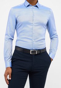 Chemise bleu clair à boutons avec une texture lisse, manches longues, col classique, et un pantalon sombre contrasté avec une ceinture noire.